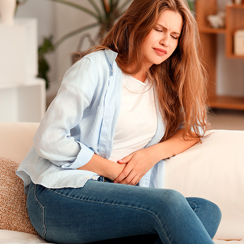 Mulher sentada no sofá com expressão de dor, segurando a barriga com as duas mãos, indicando desconforto abdominal.