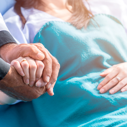 Pessoa em leito hospitalar recebendo apoio, com duas mãos entrelaçadas em gesto de cuidado e conforto.