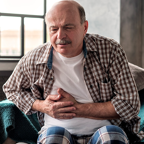 Homem sentado segura o abd&ocirc;men com express&atilde;o de dor, indicando desconforto abdominal.